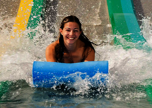 Young woman lands laughing with blue slide mat in the water at the end of a matracer water slide.