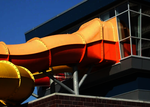 Orangefarbene Wasserrutsche mit Fassadendurchdringung verlässt Glasvorbau des Gebäudes.