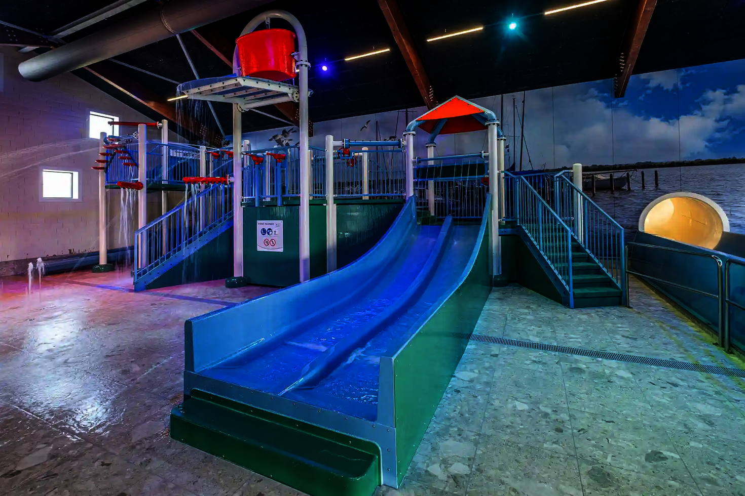 Multislide Kinderrutsche in blau und grün in einem Hallenbad, im Hintergrund weitere Wasserspielstrukturen.