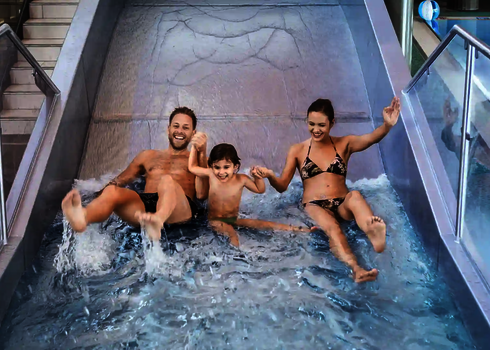 Zwei Erwachsene und ein Kind halten sich an den Händen und rutschen lachen eine Breitwellenrutsche herunter.