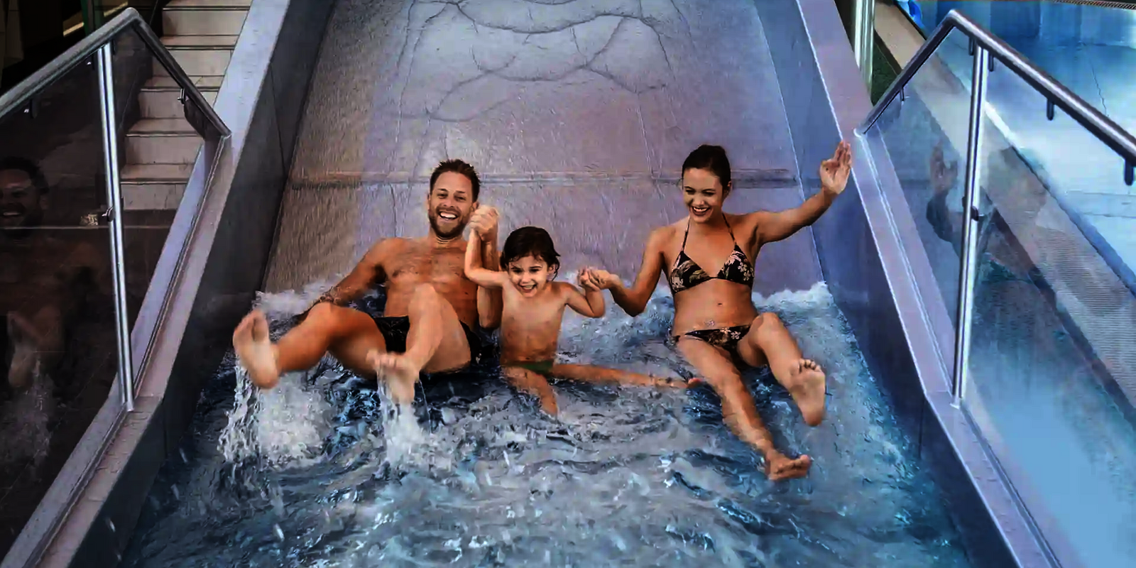 Zwei Erwachsene und ein Kind halten sich an den Händen und rutschen lachen eine Breitwellenrutsche herunter.