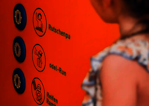 Kind steht vor oranger Wand mit beleuchteten Knöpfen und Symbolen zur Themenauswahl.
