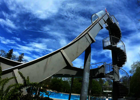 Freifallrutsche mit starkem Gefälle und Treppenturm neben einem Außenbecken.