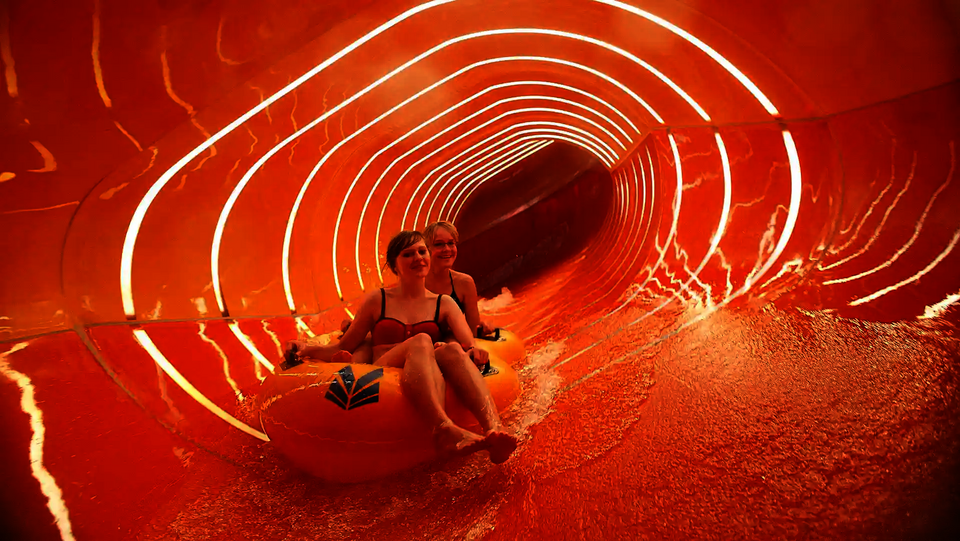 Zwei Personen rutschen auf gelbem Reifen durch eine orangene Family Twister Wasserrutsche.