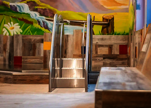  Kinderrutsche aus Edelstahl mit Treppe in einem Hallenbad, vor bunter Wandmalerei mit Blumen und Wasserfall.