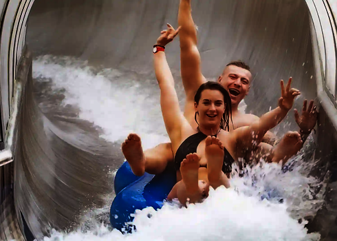 Zwei Personen in Badebekleidung rutschen lachen eine Rutsche herunter.