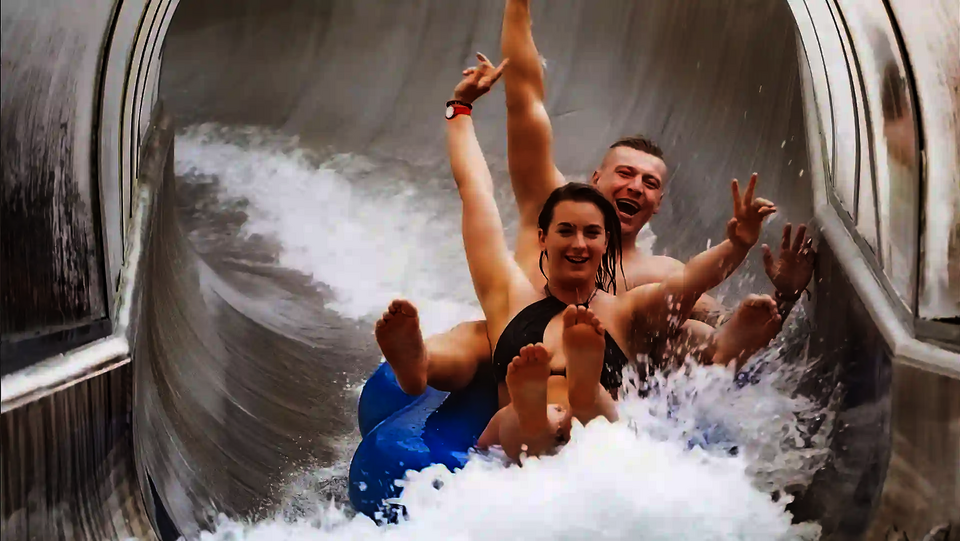 Zwei Personen in Badebekleidung rutschen lachen eine Rutsche herunter.
