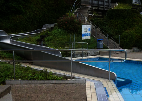Long stainless steel slide in the outdoor pool with run-outs into a swimming pool and green surroundings.