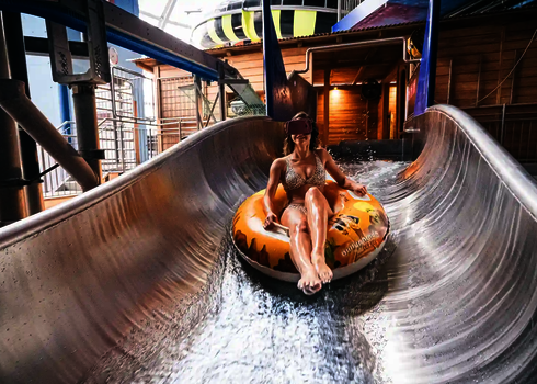 Eine Person sitzt lächelnd in einem gelben Schwimmreifen in einer Edelstahl-Wasserrutsche und trägt eine VR-Brille.