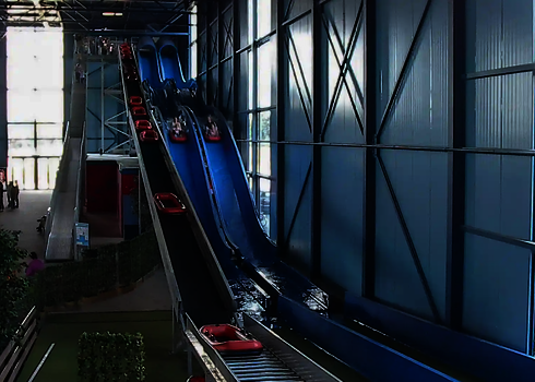 Überdachter Innenbereich eines Freizeitparks mit einer blauen Harakiri Wasserrutsche, daneben ein Reifenlift.