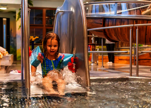  Kind mit Schwimmflügeln rutscht lachend auf Edelstahlrutsche ins flache Becken.