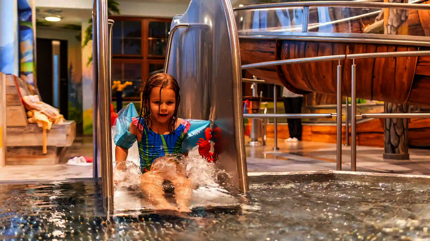  Kind mit Schwimmflügeln rutscht lachend auf Edelstahlrutsche ins flache Becken.