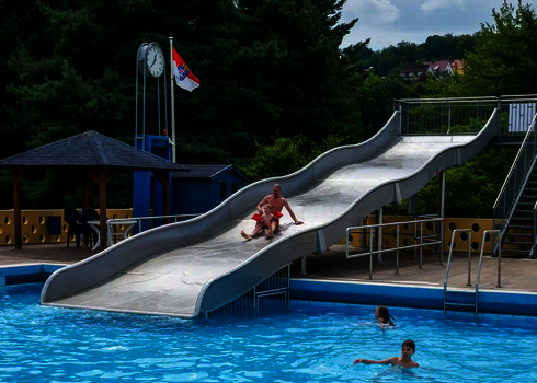 Blick auf ein Freibad mit einer Breitwellenrutsche, auf der ein Mann mit Kind rutscht.