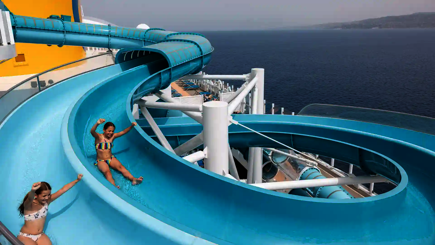 Kinder rutschen lachend die blaue 2-in-1 Racer Wasserrutsche auf einem Kreuzfahrtschiff hinunter.