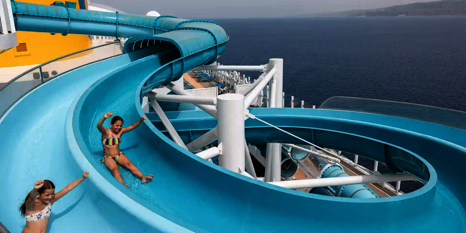 Kinder rutschen lachend die blaue 2-in-1 Racer Wasserrutsche auf einem Kreuzfahrtschiff hinunter.