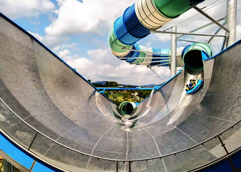 Nahaufnahme eines blauen Urknalls aus Edelstahl, Personen auf blauen Rutschreifen kommen gerade auf die Rutsche.