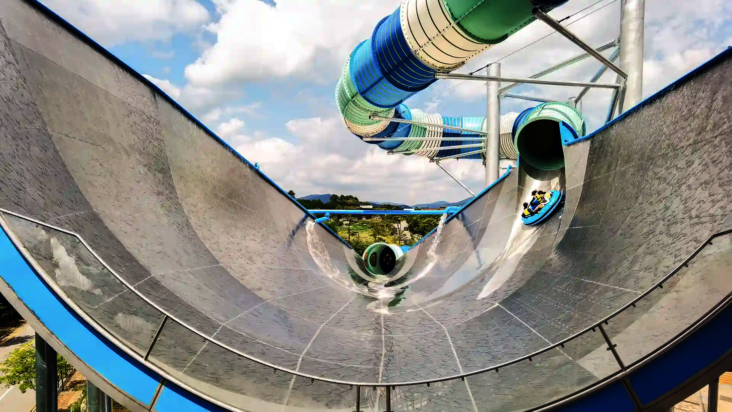 Nahaufnahme eines blauen Urknalls aus Edelstahl, Personen auf blauen Rutschreifen kommen gerade auf die Rutsche.