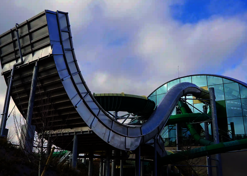 Außenansicht der Boomerang-Rutsche mit hohem Auffahrbogen vor einem Glasgebäude.