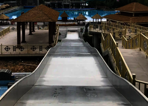 Blick von oben auf eine Breitwellenrutsche, die in einem Auslaufbecken endet, nebenan sind kleine Hütten mit Strohdächern.