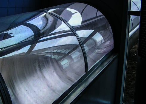 Detail einer gebogenen Wasserrutsche mit klarem Tunnel bei Fassadendurchdringung.