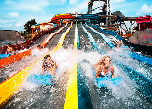 Several people slide side by side on blue mats down a colourful water slide.