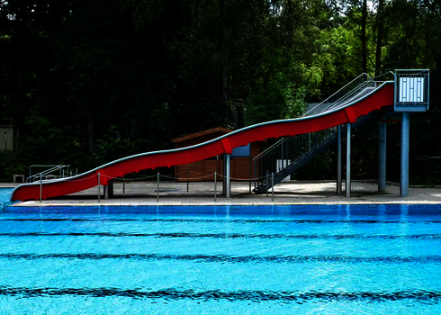Eine lange Breitwellenrutsche mit roten Seitenwänden hinter einem blauen Schwimmbecken und vor einem Wald.