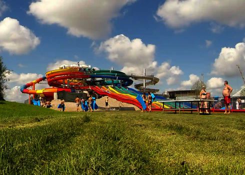 Besucher gehen mit blauen Matten auf einer Wiese zur Wasserrutsche Matracer, bunte Röhren im Hintergrund.