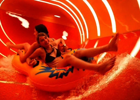 Zwei Personen rutschen lachend auf einem Doppelreifen durch den beleuchteten Family Twister.