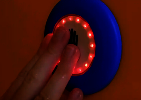 Hand berührt rot leuchtenden Auswahlknopf mit Hand-Symbol an der orangen Wand.