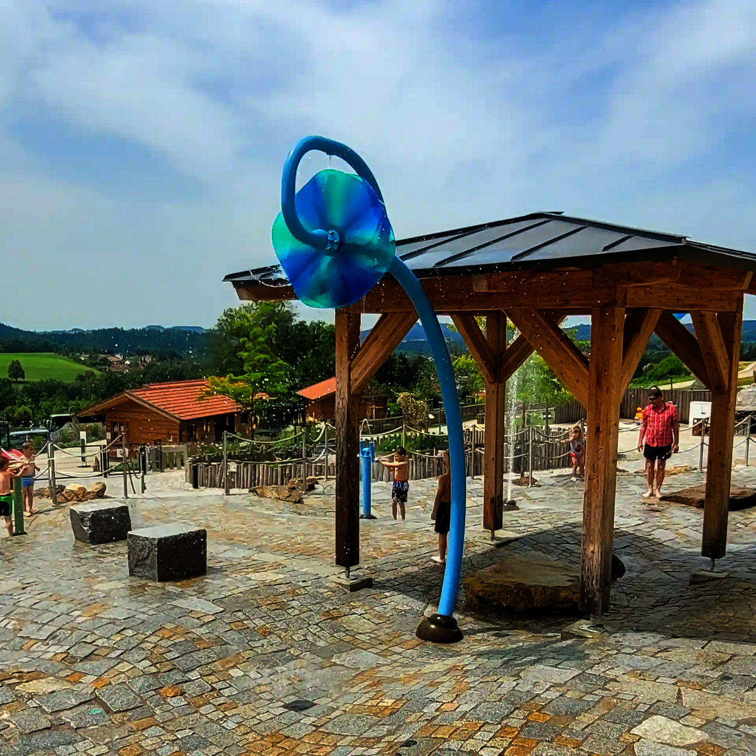 Wasserspielplatz mit Wie-Spray Element "Wasserrad" vor bergigem Hintergrund.
