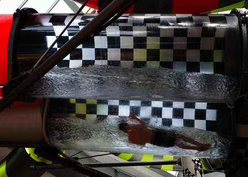 Eine Person gleitet durch eine transparente Röhre einer Wasserrutsche mit Schachbrettmuster und Wasserfluss.