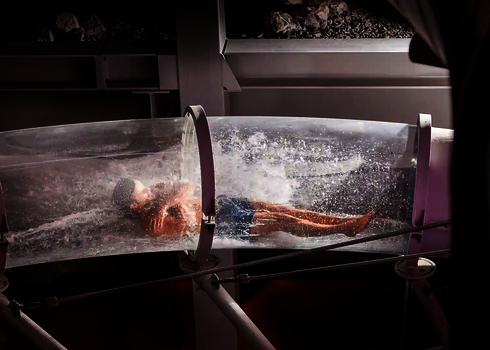 Person rutscht in durchsichtiger Röhre mit starkem Wasserfluss durch eine Indoor-Anlage.
