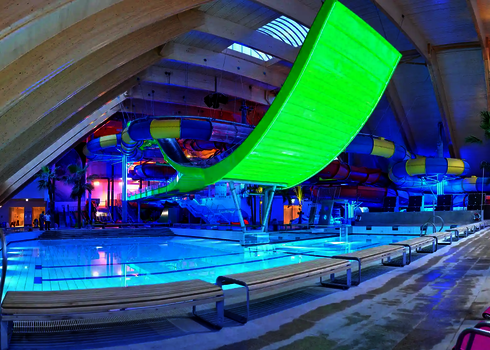 Weitwinkelansicht des Indoor-Pools mit grün leuchtender Boomerang-Rutsche und Liegen.