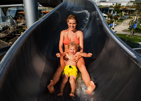 Mutter rutscht mit Kind auf Schoß eine Edelstahl-Wasserrutsche in Freizeitbad hinunter.