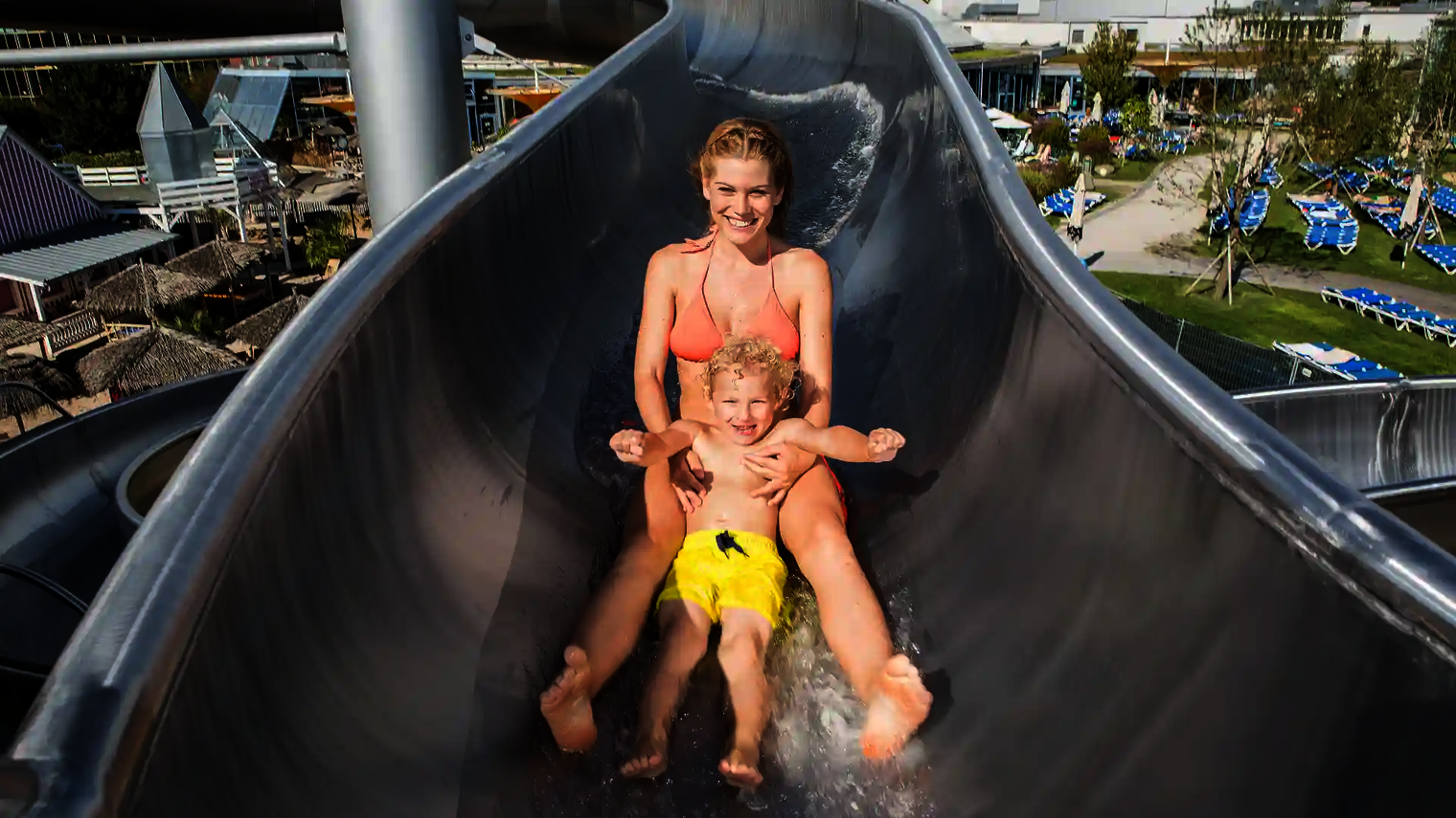 Mutter rutscht mit Kind auf Schoß eine Edelstahl-Wasserrutsche in Freizeitbad hinunter.
