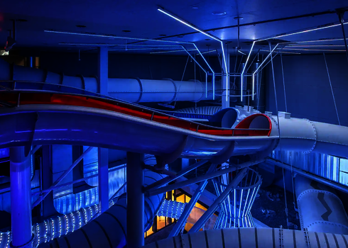 Offene Rutsche mit rotem Rand, umgeben von blau beleuchteten Röhren und Lichttrichtern.